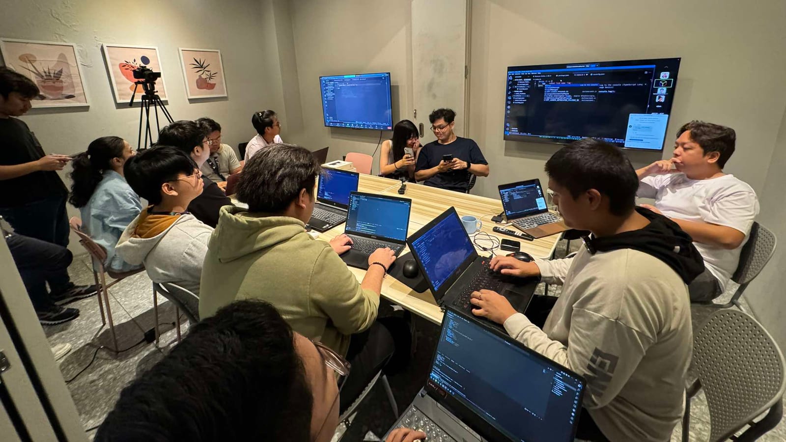 Incubation team coding side by side on laptops at a shared table with their work mirrored on twin wall monitors during a build session.