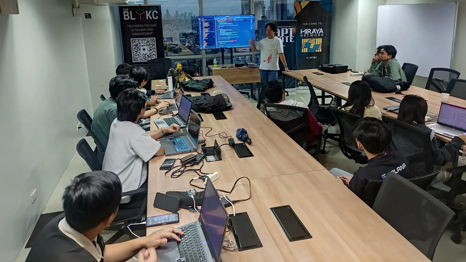 Builder presenting a live demo of code on screen to seated teammates beneath The BLOKC and Hiraya Network banners.