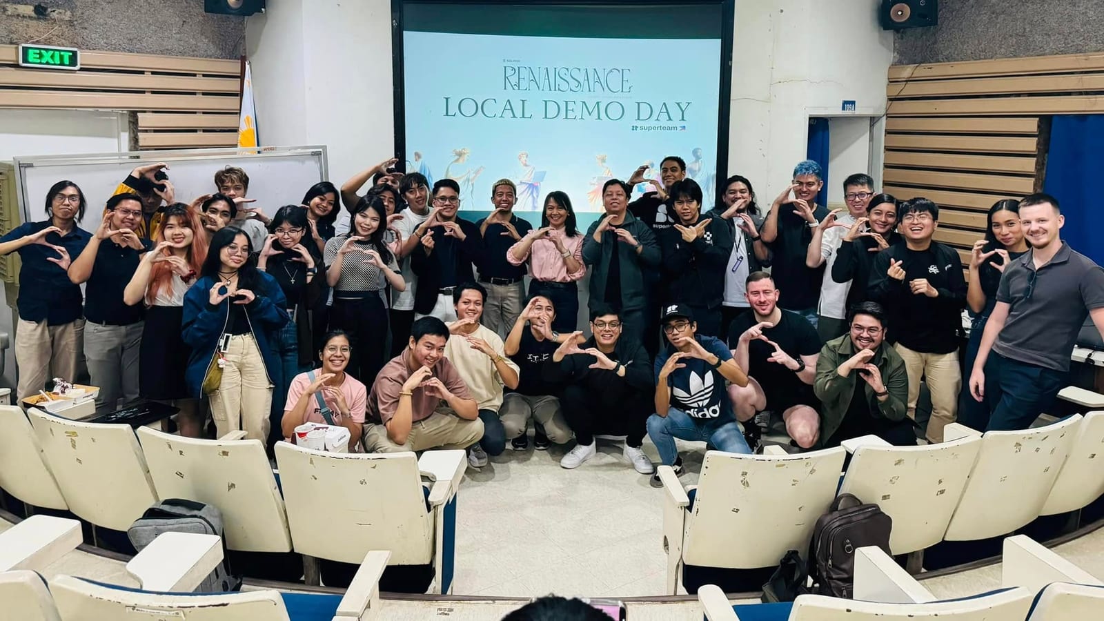 A group of participants forming heart hands together beneath the Renaissance Local Demo Day banner in an auditorium.