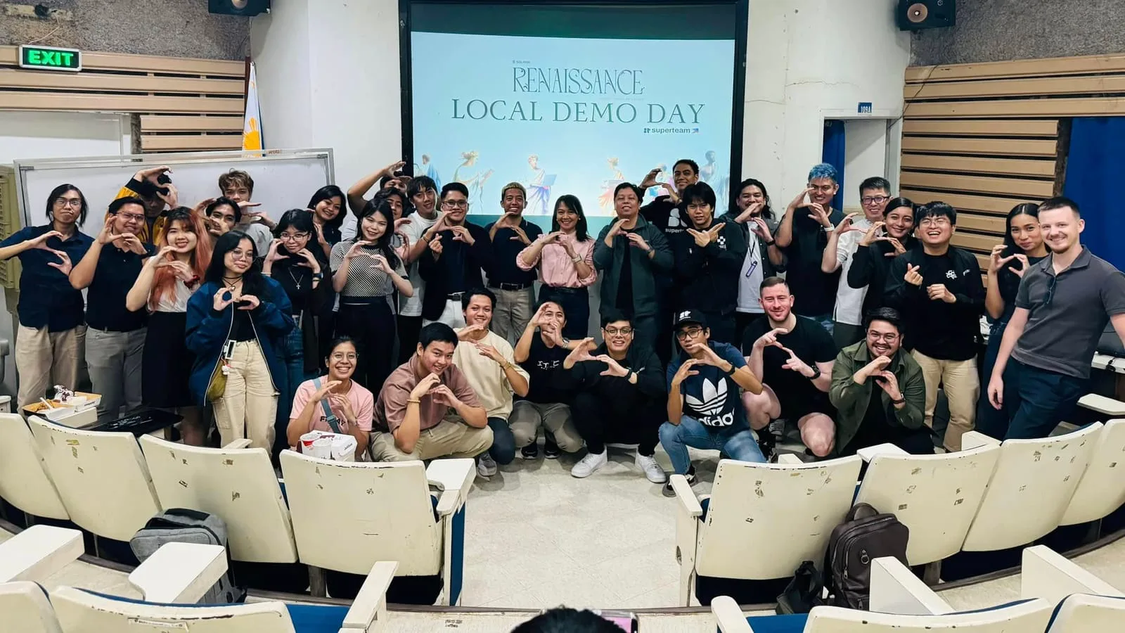 A group of participants forming heart hands together beneath the Renaissance Local Demo Day banner in an auditorium.