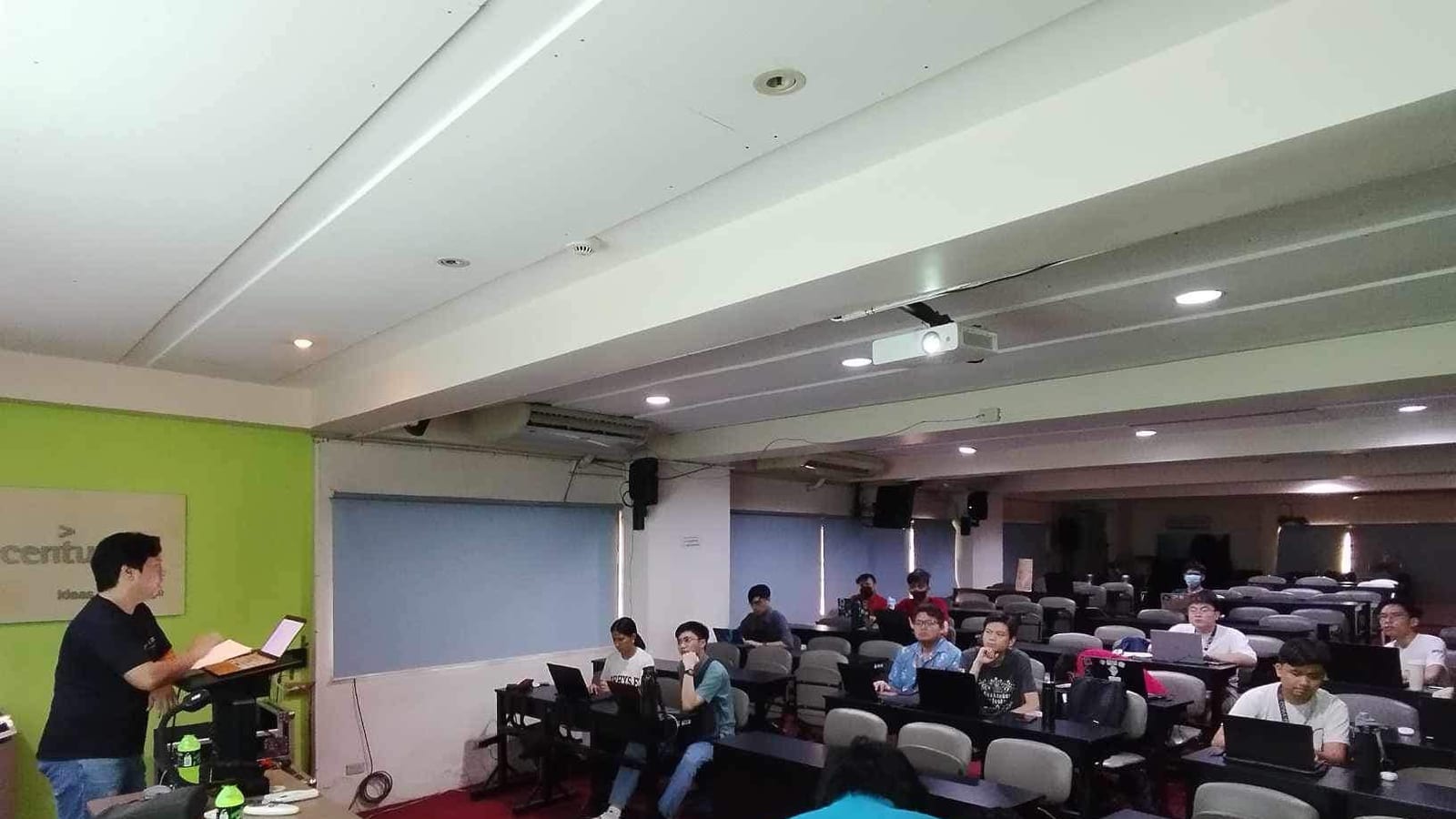 A speaker presents beside an Accenture-branded green wall to students seated at laptops in a tiered lecture room.