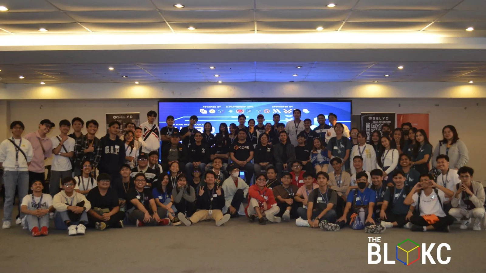 A large BLOKC bootcamp cohort gathers for a group photo in front of a blue-lit screen listing the event's powering, partner, and sponsoring Web3 organizations.