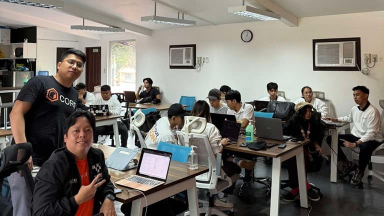 Two BLOKC organizers, one in a Core shirt, sit with participants coding at desks in a bright coworking space during a CoreDAO bootcamp.