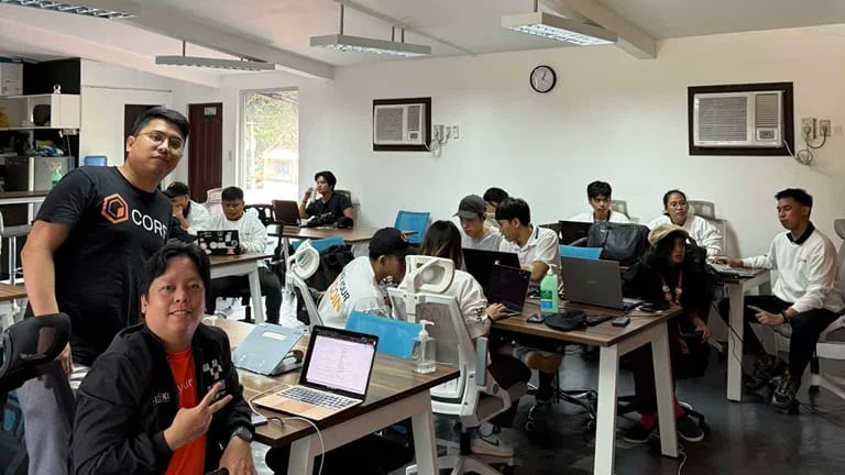 Two BLOKC organizers, one in a Core shirt, sit with participants coding at desks in a bright coworking space during a CoreDAO bootcamp.