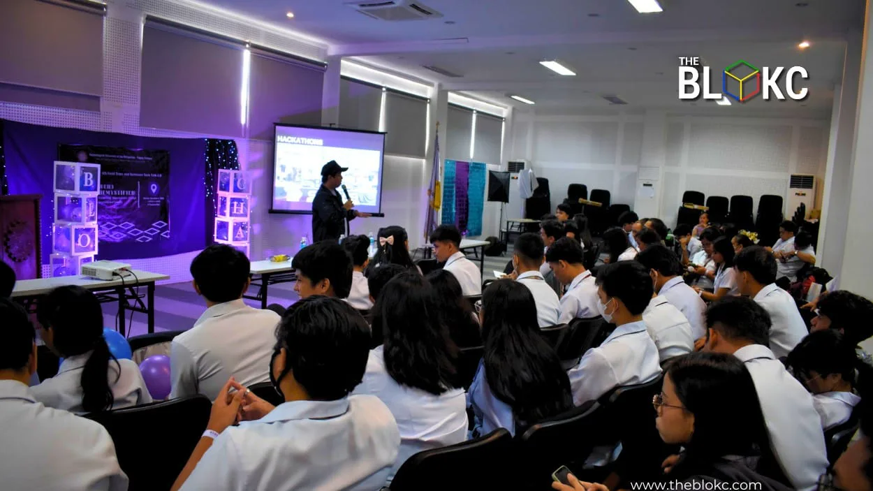 An audience watches a hackathons presentation amid BLOKC cube decorations in a purple-lit BLOKC workshop hall.