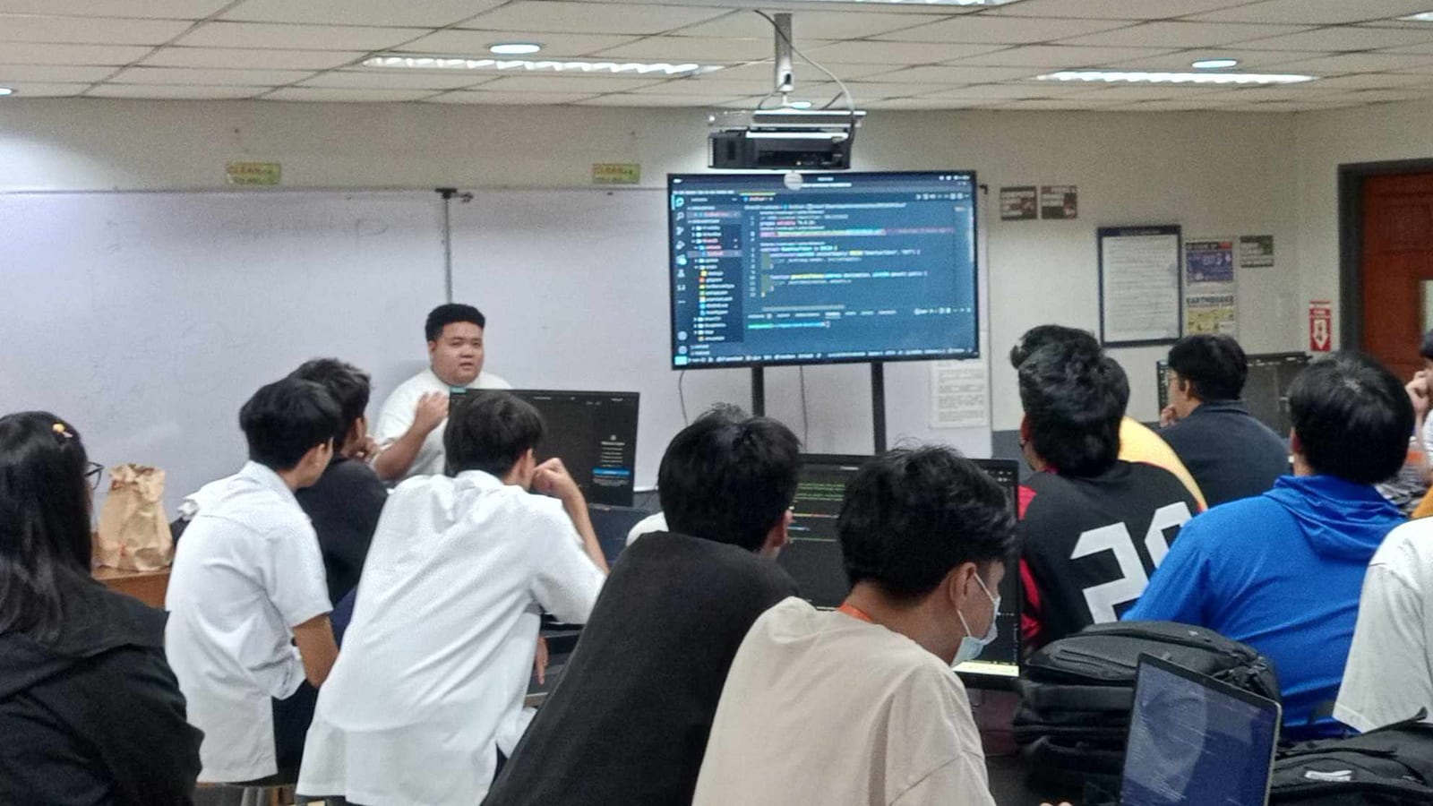 A speaker walks students through code shown on a large monitor in a university computer lab during a BLOKC developer bootcamp.