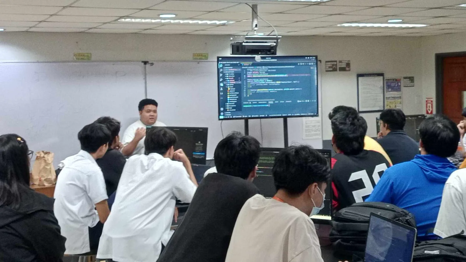 A speaker walks students through code shown on a large monitor in a university computer lab during a BLOKC developer bootcamp.