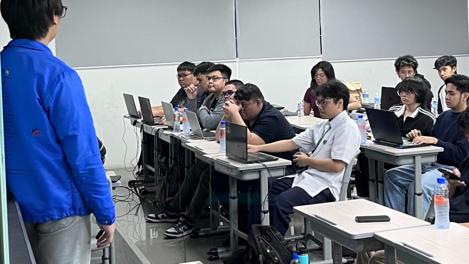 A speaker in a blue jacket addresses students working on laptops at desks in a modern BLOKC bootcamp classroom.