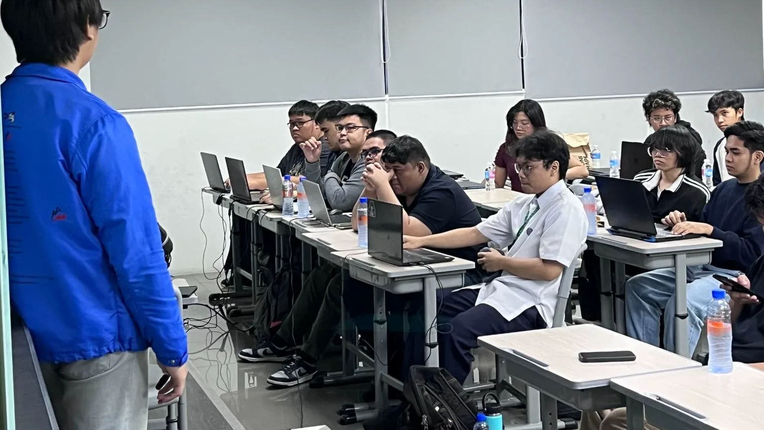 A speaker in a blue jacket addresses students working on laptops at desks in a modern BLOKC bootcamp classroom.