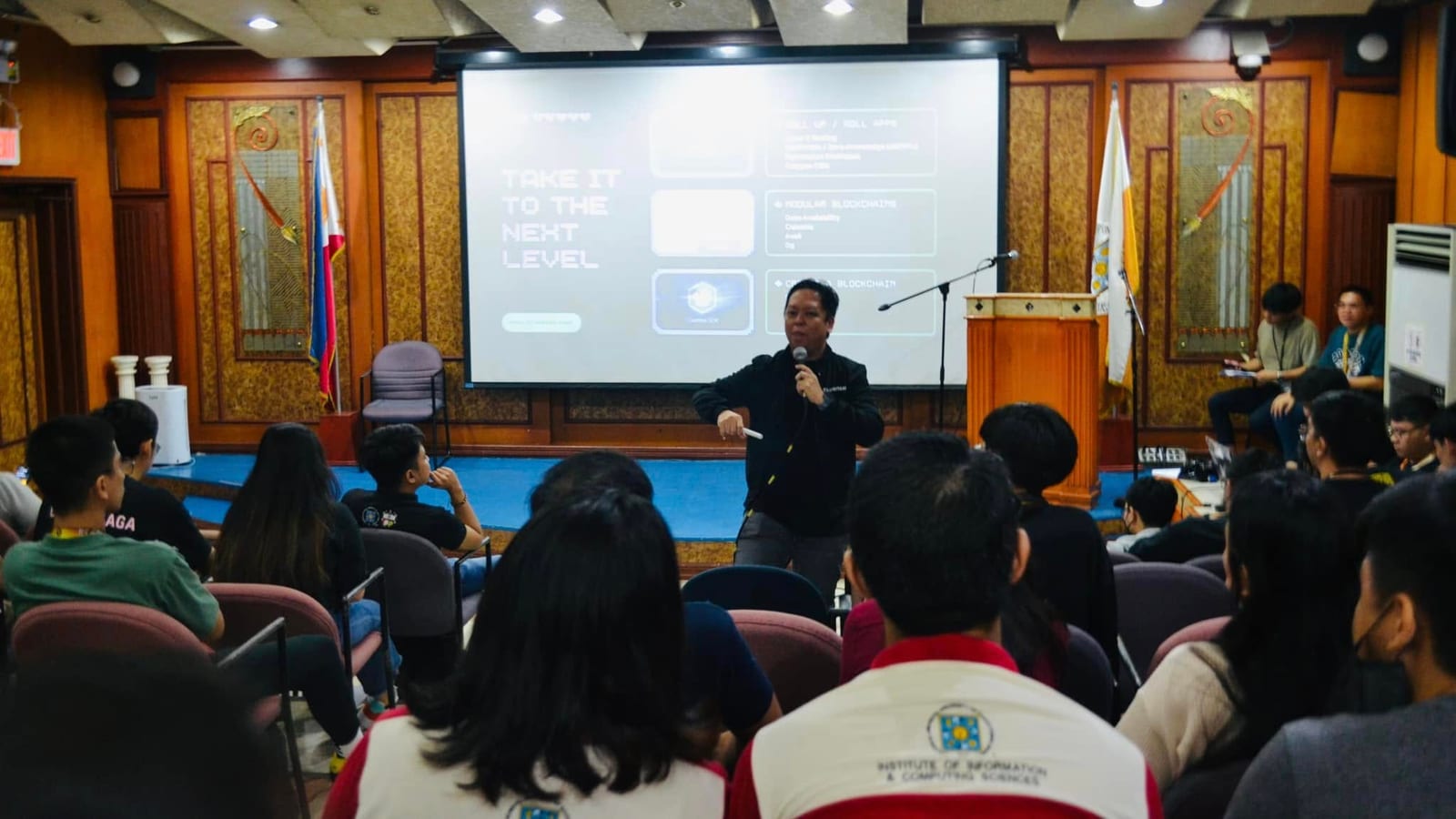 A speaker presents a Social Media session to students seated at desktop computers in a blue-and-yellow university lab.