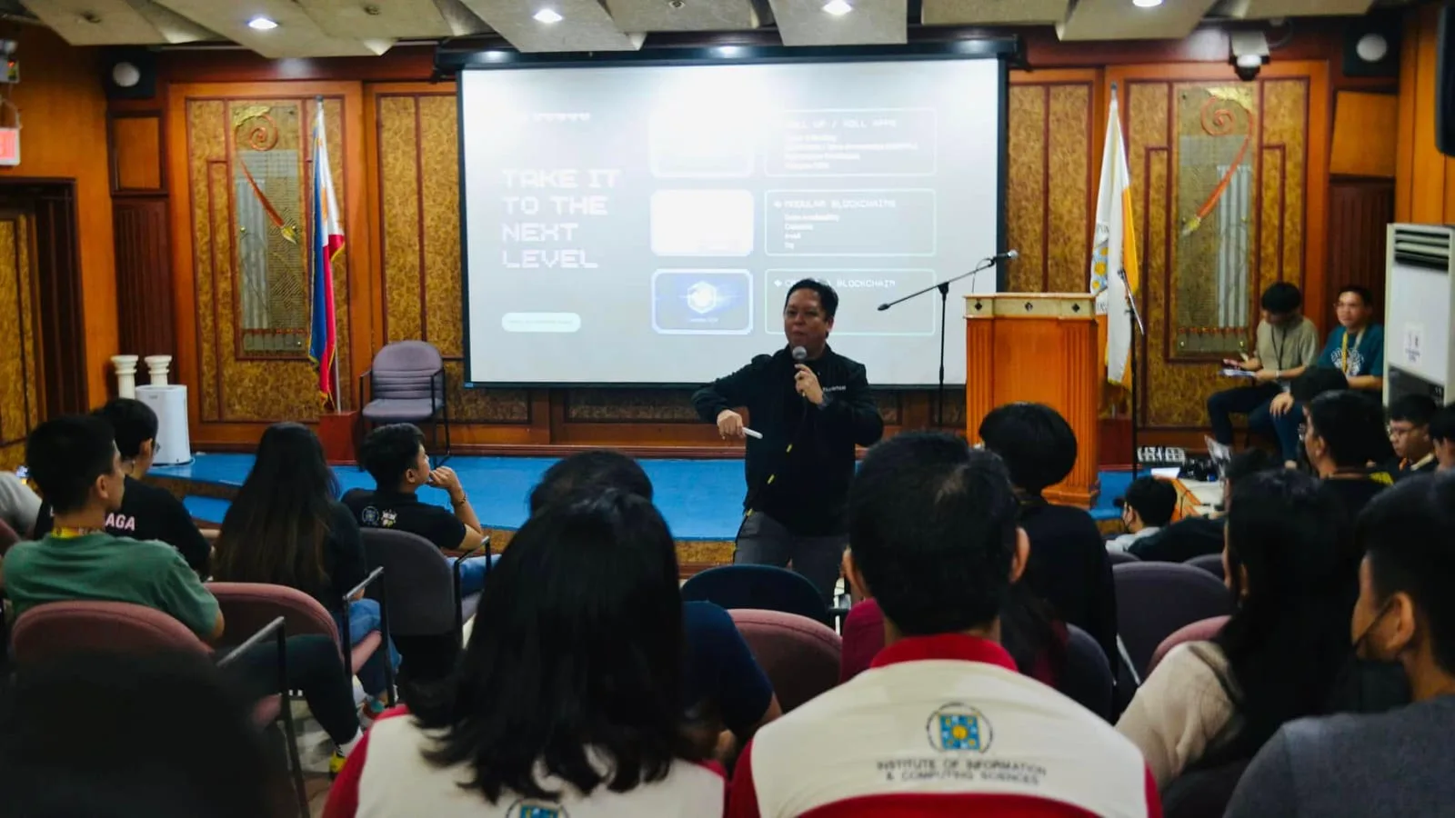 A speaker presents a Social Media session to students seated at desktop computers in a blue-and-yellow university lab.
