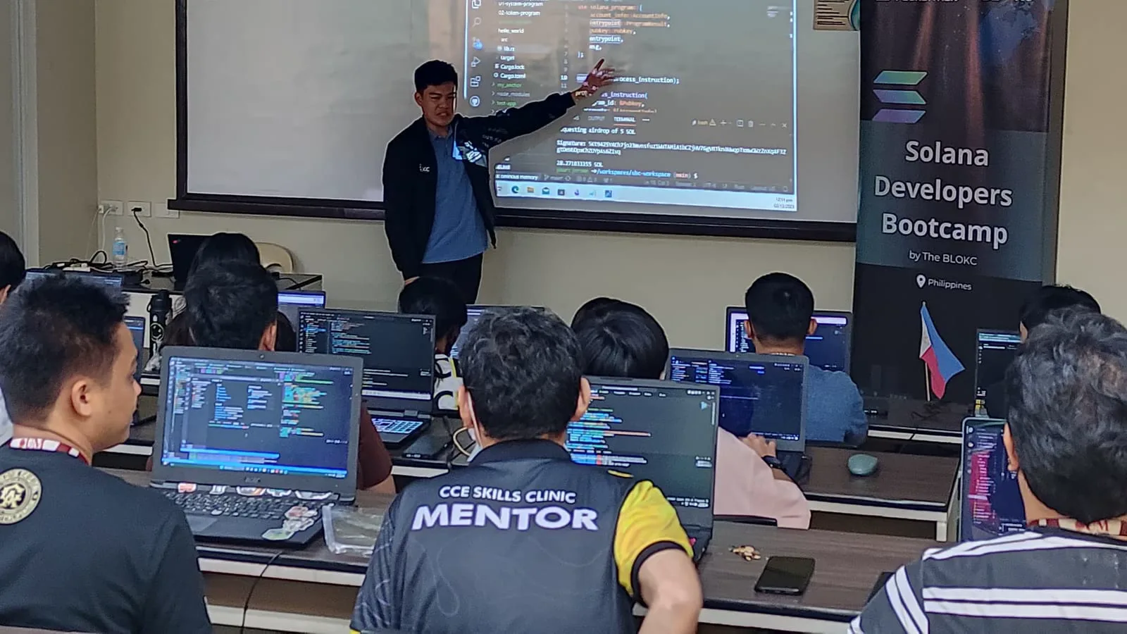 An instructor points to live Solana code on screen at The BLOKC's Solana Developers Bootcamp in the Philippines while students follow along on laptops.