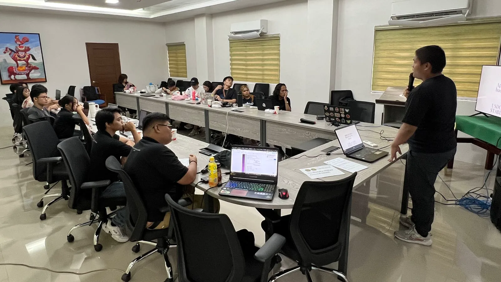 A speaker with a microphone presents a BLOKC training session to participants seated with laptops around a long conference table.